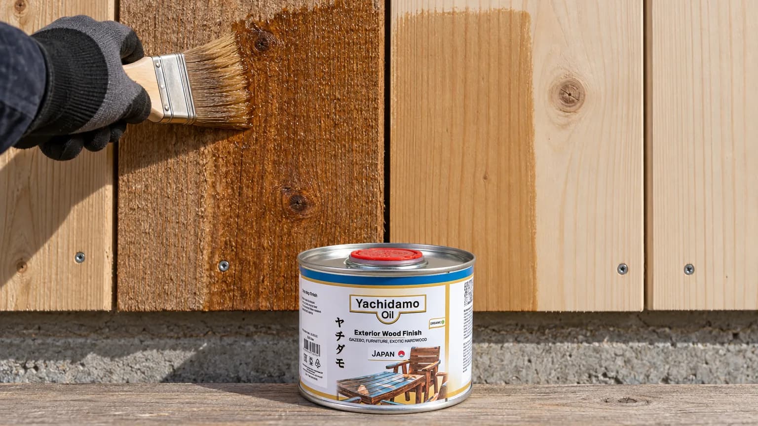 Банка Yachidamo Oil Exterior Wood Finish стоит у фасада с тремя тестовыми полосами разной степени впитывания масла.