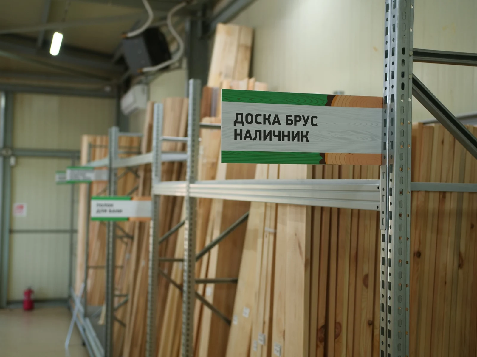 Складской зал: стеллажи с вертикальными пачками погонажа, указатель «Доска брус наличник», огнетушитель у стены
